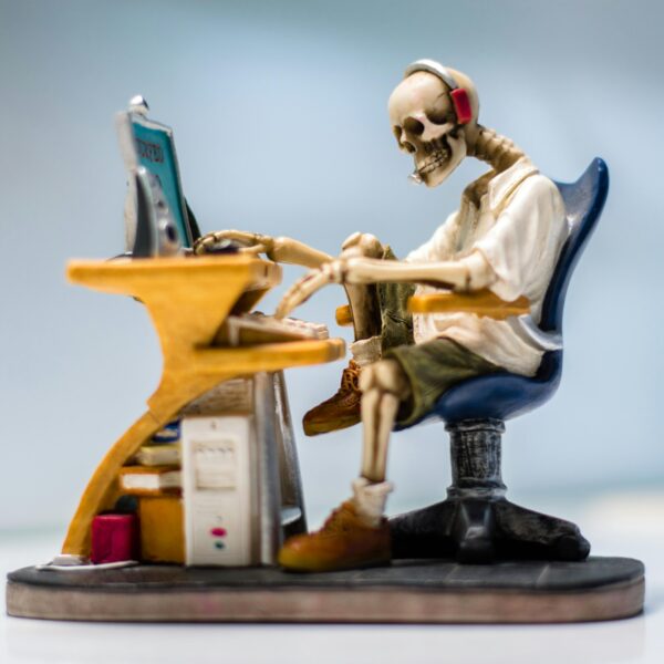 A quirky skeleton figurine wearing headphones sits at a desk with a computer, symbolizing burnout.